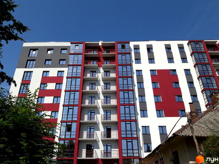 Фасад багатоповерхового будинку Житлового комплексу Центральний з білими, червоними та сірими секціями. Навколо зелені дерева та стара забудова.