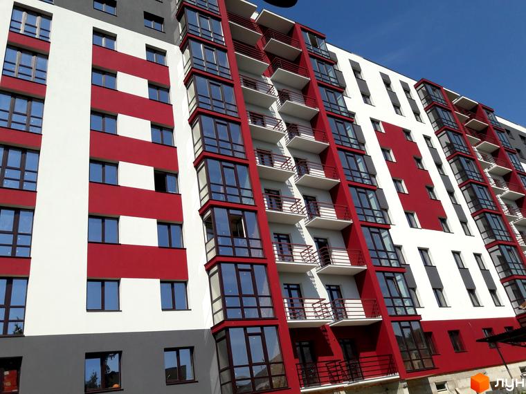 Зовнішній вигляд Житлового комплексу Центральний: багатоповерхова будівля з червоно-білим фасадом та великими вікнами. Навколо будинку облаштована територія.