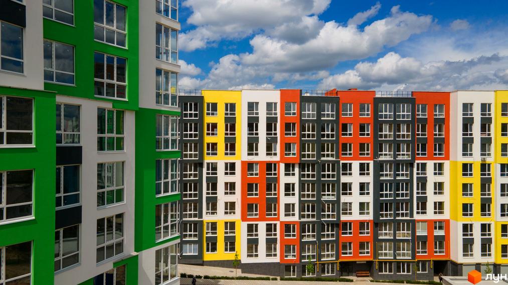 Зовнішній вигляд фасаду житлового комплексу Синергія-3+ з яскравими кольоровими секціями та великими вікнами. Навколо будинку облаштована прибудинкова територія з зеленими насадженнями.