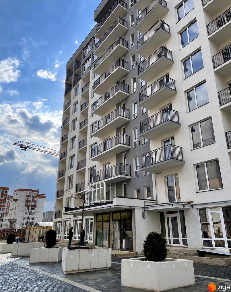 Зовнішній вигляд новобудови ЖК KromaxBud з балконами та вікнами на фоні неба з хмарами. Прибудинкова територія з бетонними клумбами та деревами.