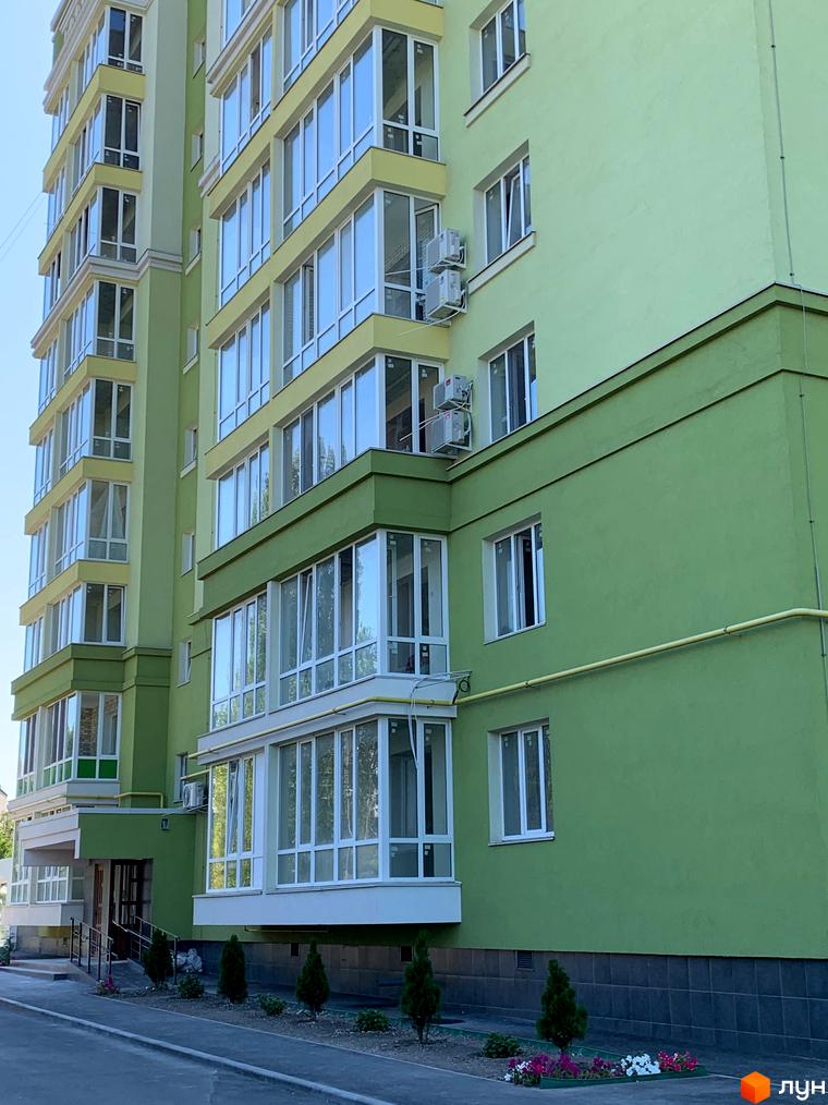 Зовнішній вигляд фасаду світло-зеленої новобудови ЖК Levanevsky з великою кількістю скляних балконів. Біля будинку висаджені молоді дерева та квіти.