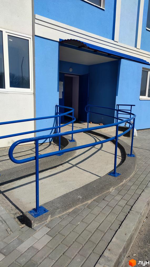 Фасад Житлового комплексу Пролісок у синьо-білих кольорах з входом та пандусом. Прибудинкова територія вимощена бруківкою.