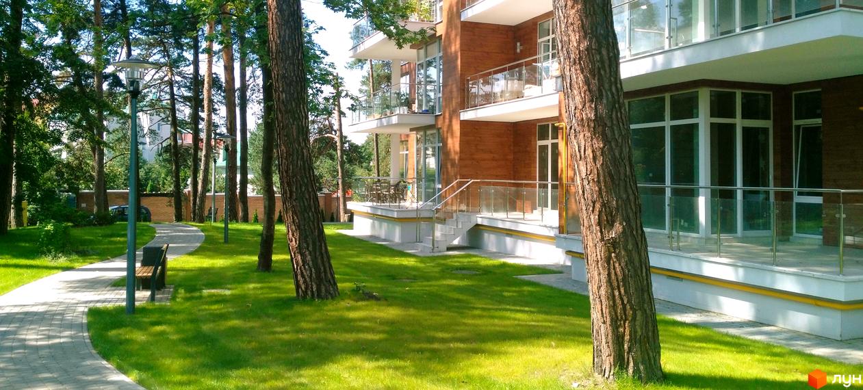 Житловий комплекс Грінвуд: фасад новобудови з дерев'яним оздобленням та скляними балконами. Прибудинкова територія з доглянутим газоном, доріжками та освітленням.