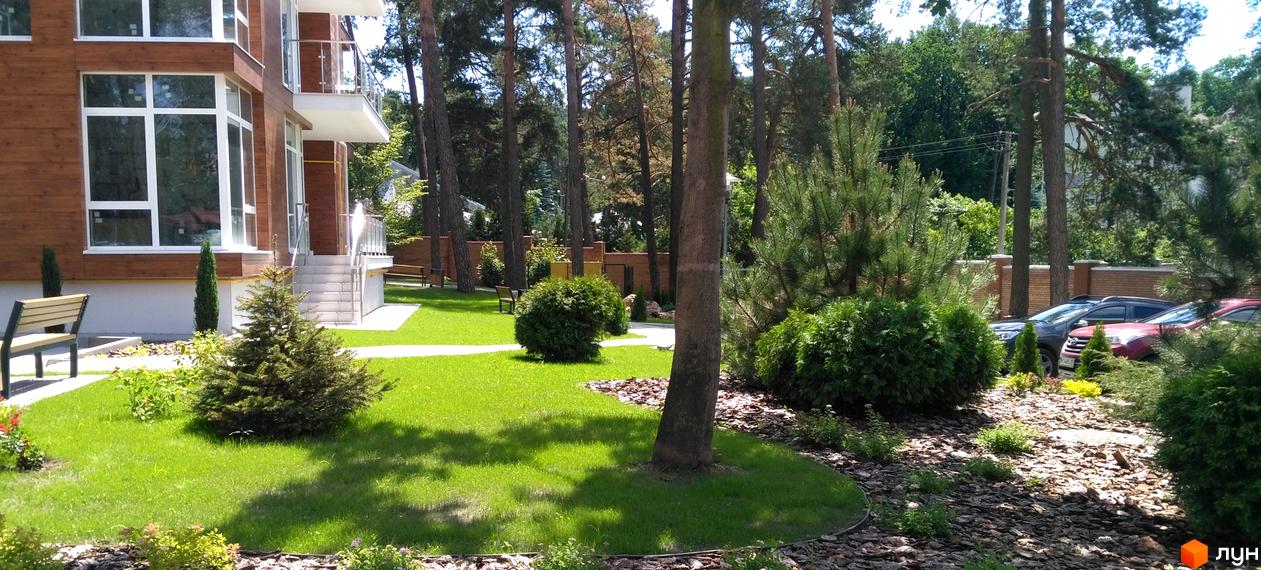 Житловий комплекс Грінвуд з дерев'яними фасадами та великими вікнами. Навколо зелений газон, дерева та місця для відпочинку.