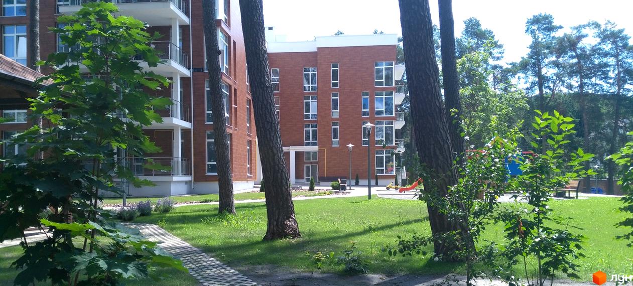 Новобудова ЖК Грінвуд з цегляним фасадом та балконами. Прибудинкова територія з зеленим газоном, деревами та дитячим майданчиком.