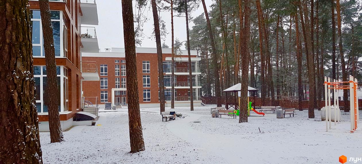 ЖК Грінвуд: цегляний багатоповерховий будинок з великими вікнами та балконами. Навколо засніжений двір з соснами, дитячим майданчиком та альтанкою.