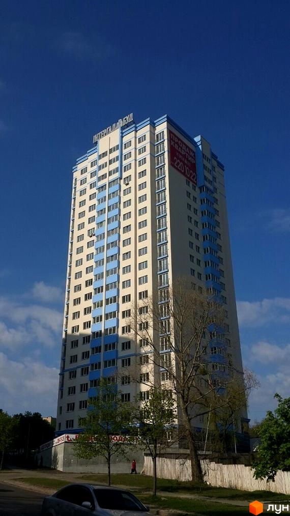 Висотна новобудова ЖК Деміївка з блакитними елементами фасаду та великою кількістю вікон. На будівлі видно рекламу продажу квартир. Навколо будівлі дерева та невелика прибудинкова територія.