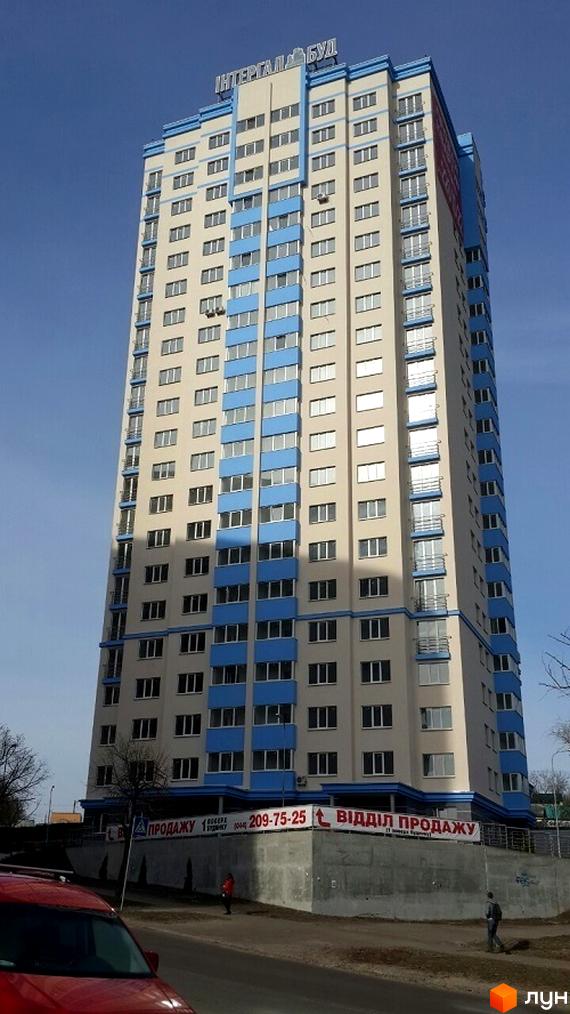 Високоповерховий житловий комплекс з блакитними елементами на фасаді та великою кількістю вікон. На першому поверсі видно банер з написом 