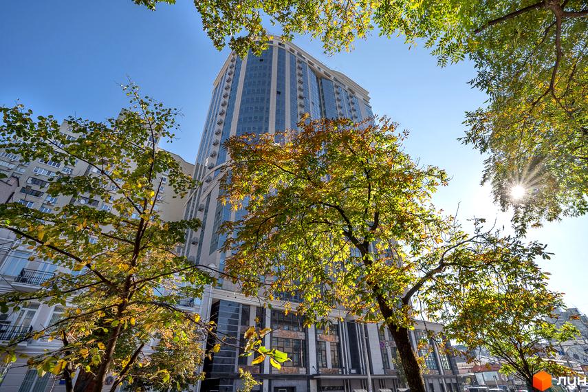 Висотна будівля ЖК Володимирський з великими вікнами, що відбивають небо. На передньому плані дерева з жовтим листям, що додають природний акцент.