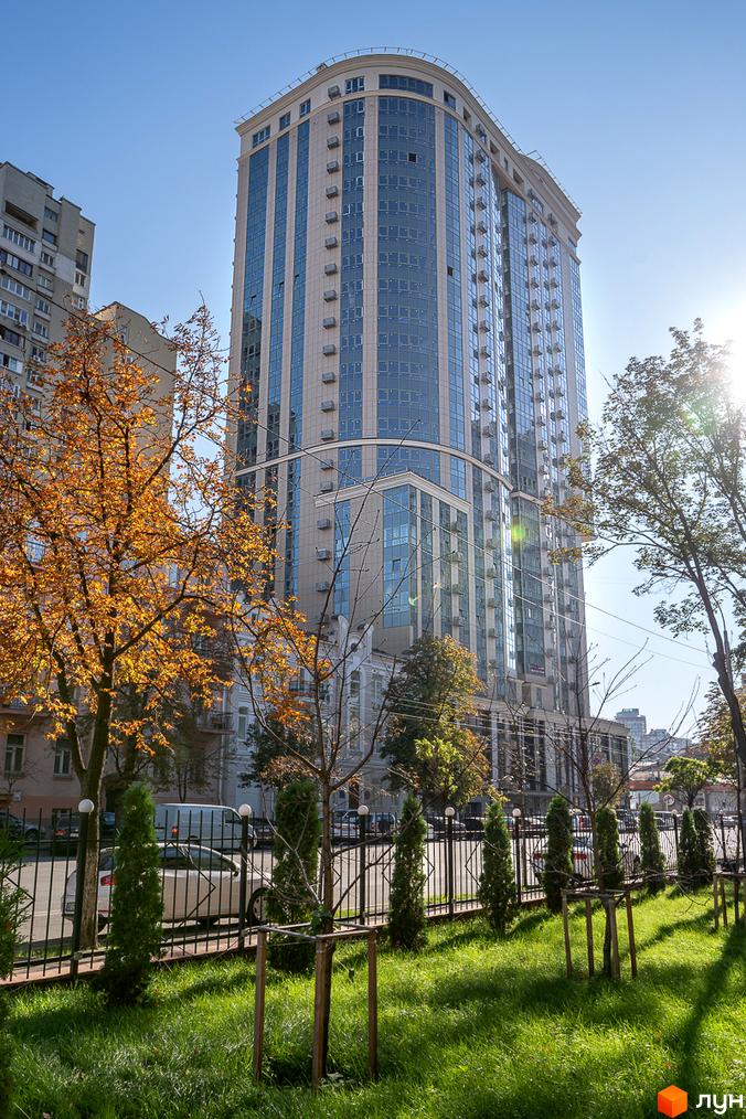 Висотна будівля житлового комплексу Володимирський з великою кількістю вікон. На передньому плані зелена трава, дерева та ковані огорожі.