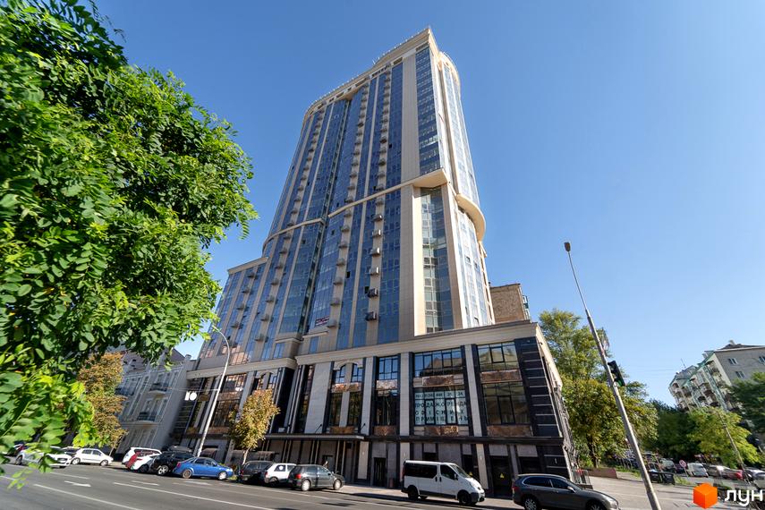 Висотна будівля ЖК Володимирський з великою кількістю вікон. На передньому плані дорога з припаркованими автомобілями та зелені насадження.