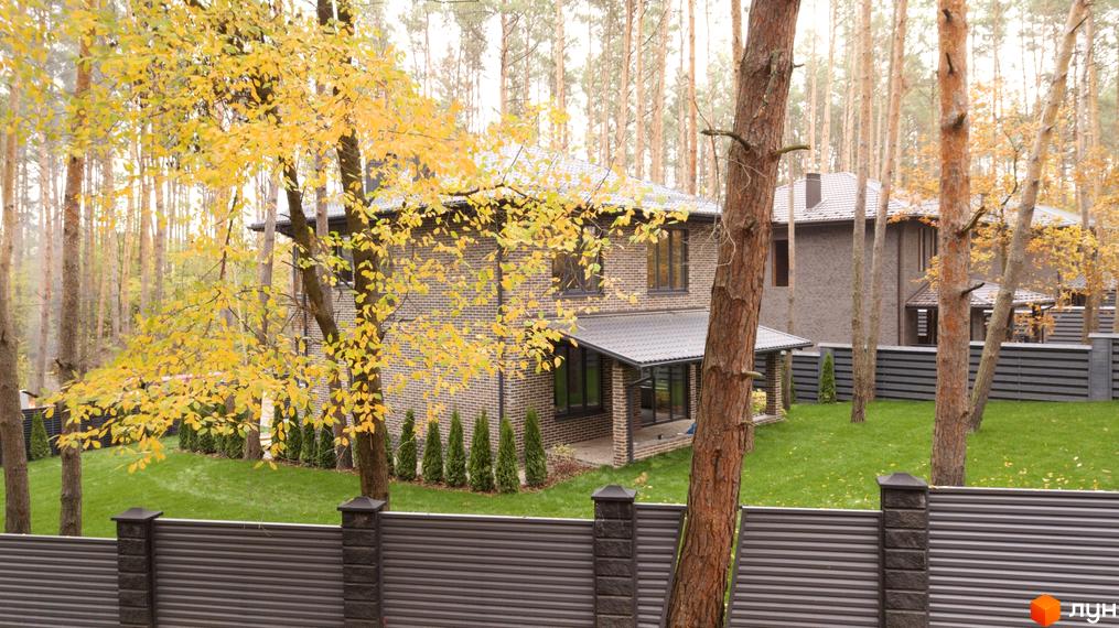 Двоповерховий будинок з коричневої цегли з темною покрівлею та великими вікнами. На території Містечка котеджів Квітка Полонини зелена галявина та дерева.