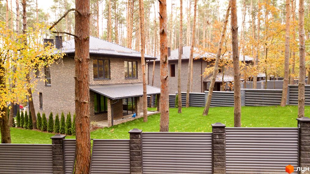 Двоповерховий будинок з цегляними стінами та темним дахом в Котеджному містечку Квітка Полонини. Зелена галявина та паркан з горизонтальними ламелями навколо будинку.