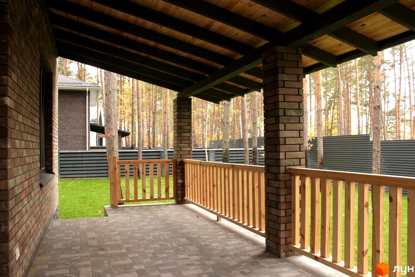 Затишна тераса будинку з цегляними колонами, дерев'яними перилами та стелею. Навколо зелена галявина та ліс, що створює атмосферу спокою.