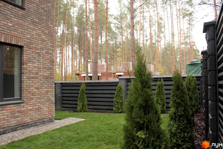 Цегляний будинок з великими вікнами, зелена галявина з туями та паркан з горизонтальними дошками. На задньому плані видно ліс.