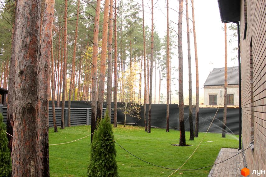 Цегляний будинок з газоном та деревами на території. На задньому плані видно Котеджне містечко Квітка Полонини.