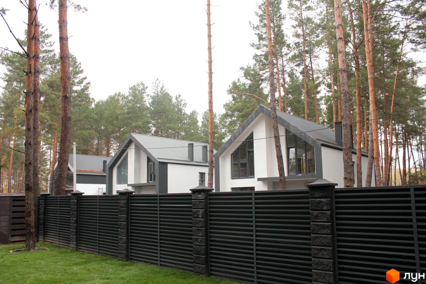 Котеджне містечко Квітка Полонини. Білі будинки з сірими дахами за чорним парканом, навколо дерева та зелена трава.