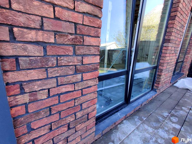 Фасад Котеджного містечка Одноповерховий Київ - Мархалівка з червоної цегли та великими вікнами. Поруч облаштована тераса з дерев'яним настилом.