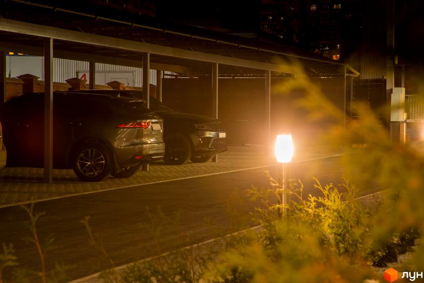 Фото парковки з автомобілями у Житловому комплексі Cофія в темну пору доби. На передньому плані декоративні кущі та ліхтар.