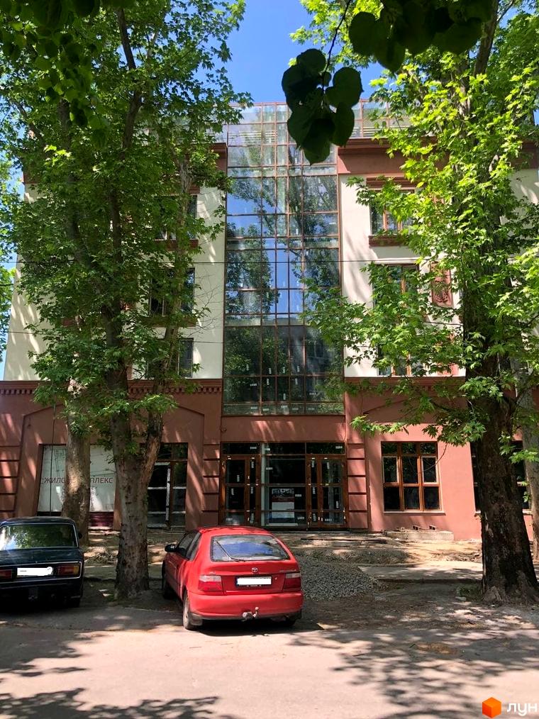 Фасад будівлі Житловий комплекс L7 House з великими вікнами та вхідними дверима. Перед будинком припарковані автомобілі, росте трава та дерева.