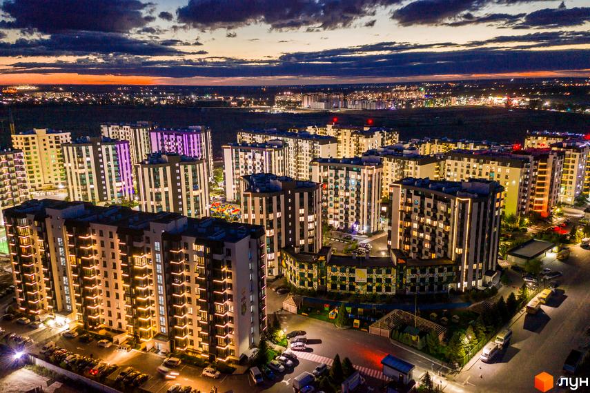 Нічний вид на житловий комплекс У-Квартал з підсвіченими фасадами будівель. Навколо комплексу розташовані дороги з автомобілями та зелені зони.