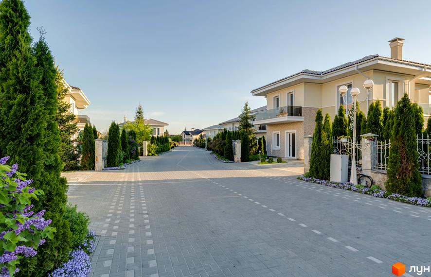 Будинок в Котеджному містечку Золоче Прованс з фасадом у світлих тонах та доглянутою прибудинковою територією з зеленими насадженнями та бруківкою.