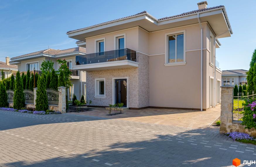 Двоповерховий будинок з бежевими стінами та скляним балконом в котеджному містечку КМ Золоче Прованс. Бруківка перед будинком та зелені насадження.