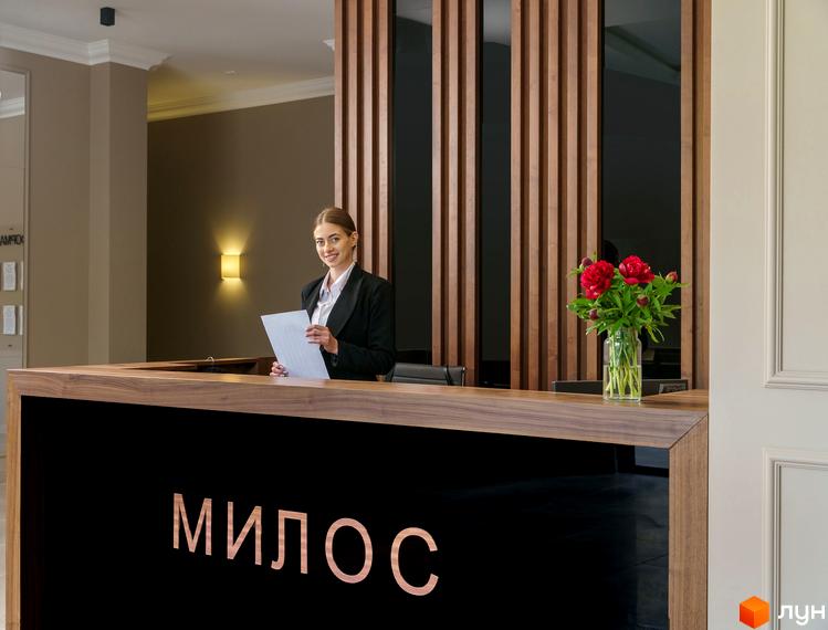 Житловий комплекс Мілос. Світлий хол з рецепцією, де працівниця зустрічає гостей. На столі стоїть ваза з квітами, позаду дерев'яні панелі.