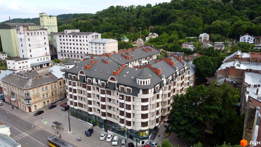 Житловий комплекс Львівський дворик має вигнутий фасад з балконами та темним дахом. Навколо – вулиця з транспортом, старі будівлі та зелений пагорб.