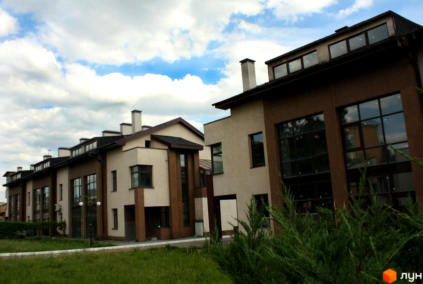 Зображення житлового комплексу. На фото представлені будинки з великими вікнами та світлими фасадами. Перед будинками зелена трава та кущі.