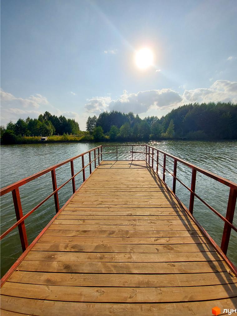Дерев'яний пірс з червоними перилами, що веде до води. На задньому плані видно ліс та сонце на небі.