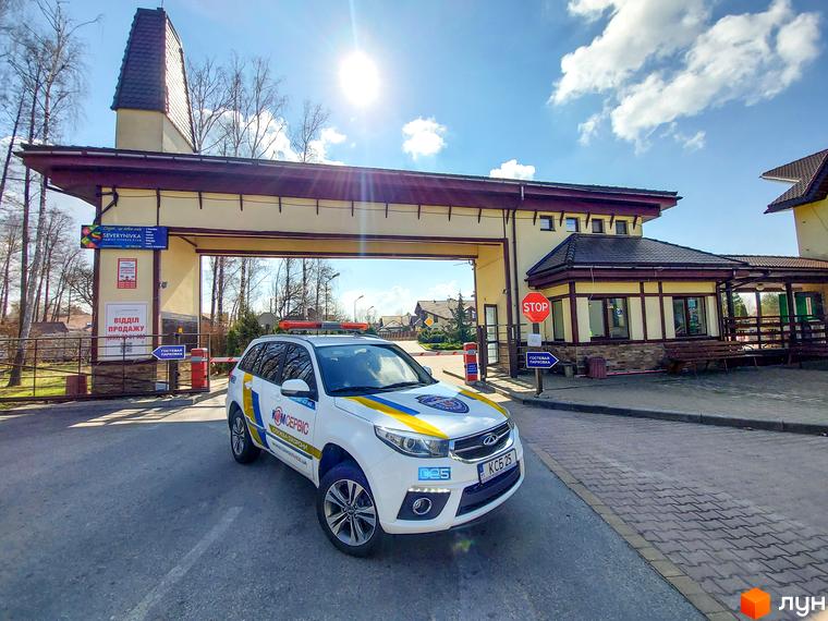 В'їзд до Містечко котеджів Северинівка. Білий автомобіль на фоні будівлі з аркою та шлагбаумом. На території бруківка та зелені насадження.