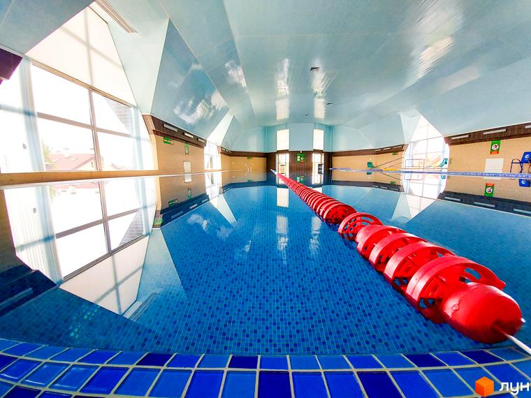 Критий басейн в Містечко котеджів КМ Северинівка. Просторий басейн з блакитною плиткою та червоною розміткою для плавання, розташований в середині приміщення з великими вікнами.