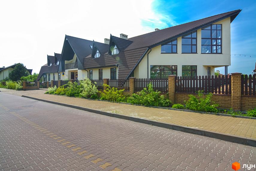 Зображення житлового будинку з похилим дахом та великими вікнами. Перед будинком розташована доглянута територія з рослинністю та бруківкою.