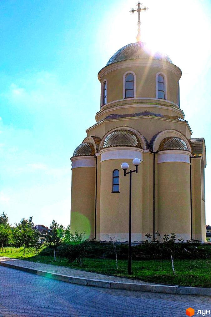 Вид на церкву з куполом та хрестом на фоні блакитного неба. Поруч розташована прибудинкова територія з газоном, деревами та тротуаром.