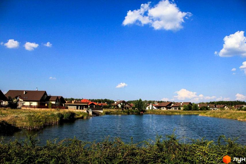 Котеджне містечко Северинівка. Мальовничий краєвид з будинками з коричневими дахами біля озера. Навколо зелена рослинність та блакитне небо з хмарами.