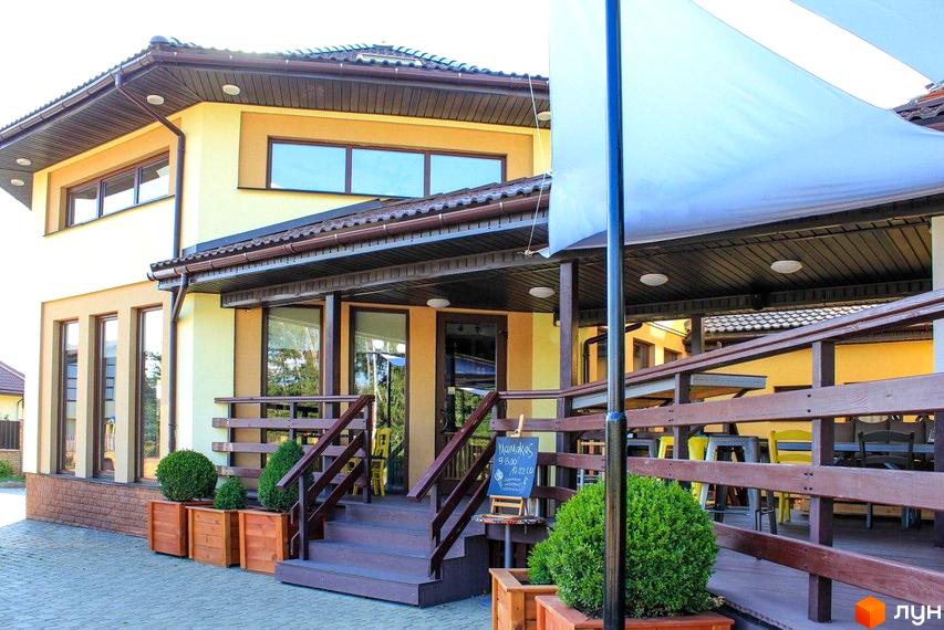 Зовнішній вигляд будинку з жовтими стінами та коричневим дахом. На передньому плані розташовані дерев'яні сходи та тераса з зеленими кущами в горщиках.