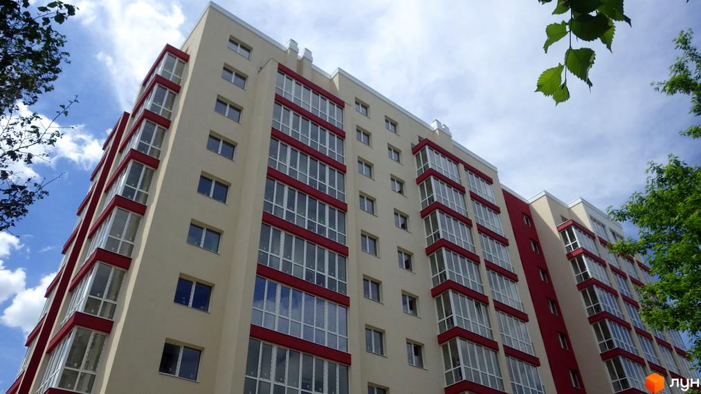 Житловий комплекс Мрія: багатоповерховий будинок світлого кольору з червоними балконами та скляними лоджіями на тлі хмарного неба та зелених дерев.