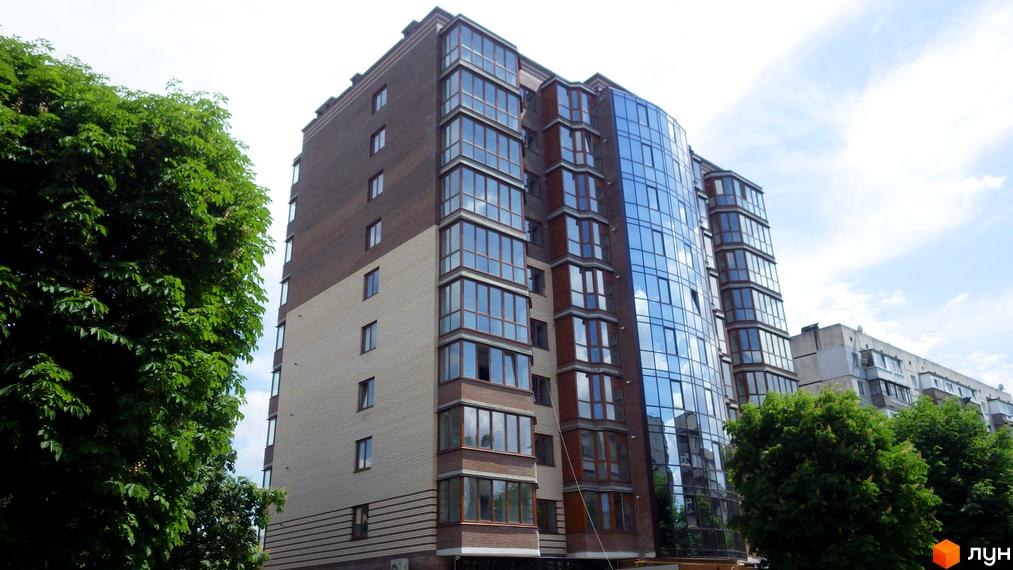 Житловий комплекс Аристократ з фасадом з коричневої та світлої цегли, великими скляними вікнами та балконами. Навколо будинку ростуть зелені дерева.