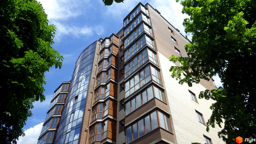 Новобудова Аристократ з цегляними стінами та панорамними вікнами, що відбивають небо, у оточенні пишних зелених дерев.