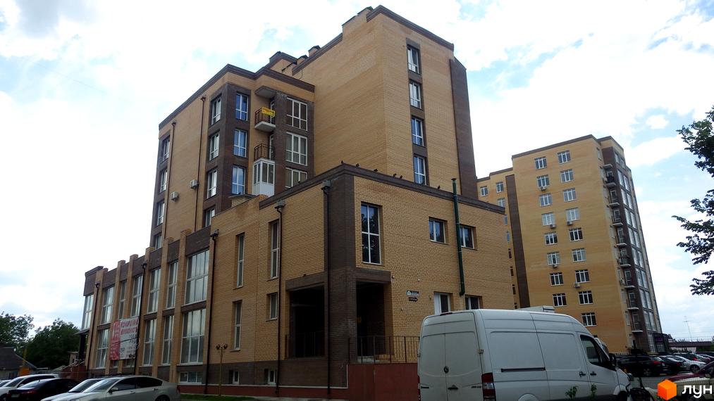 Зовнішній вигляд житлового комплексу Зелений з цегляним фасадом та численними вікнами. На прибудинковій території видно припарковані автомобілі та білий фургон.