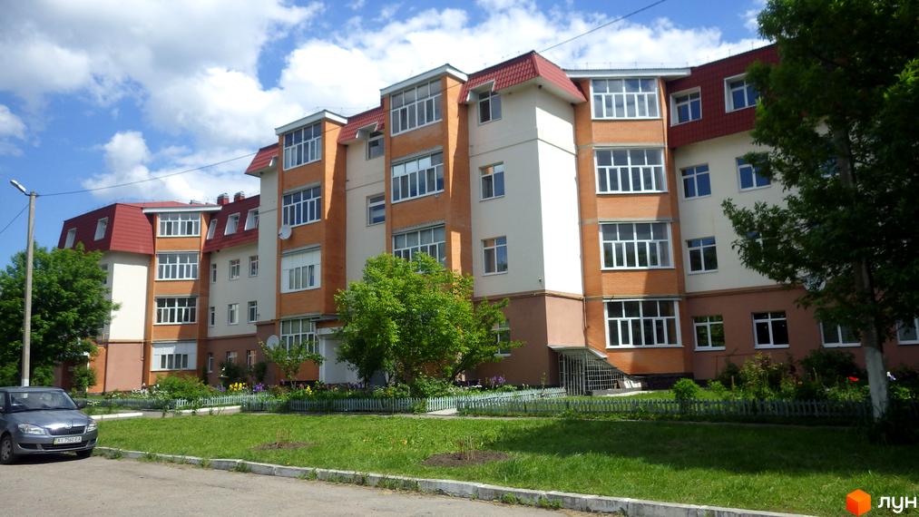 Зовнішній вигляд житлового комплексу на вул. Гайок, 224б, з цегляним фасадом та червоним дахом. Прибудинкова територія озеленена, з квітами та деревами.