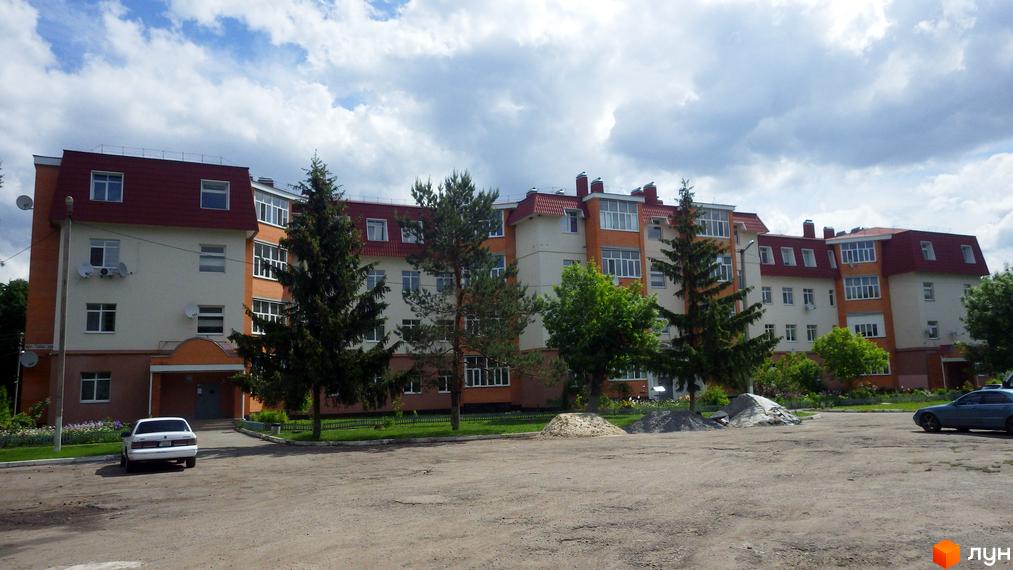 Житловий комплекс на вул. Гайок, 224б з червоним дахом та світлими фасадами. Перед будинком зелені дерева та парковка.