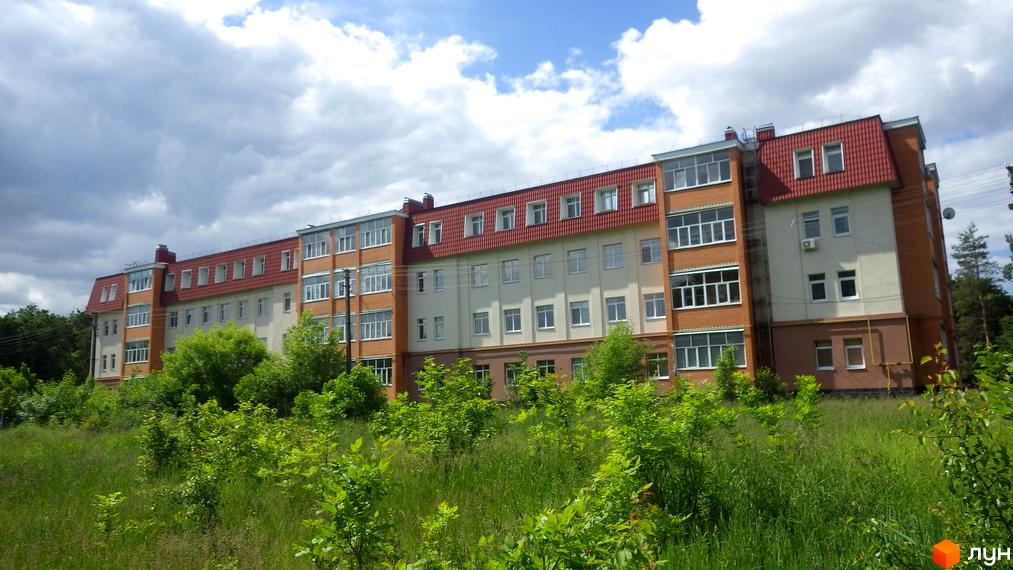 Багатоповерховий житловий будинок з червоним дахом та світлим фасадом, оточений зеленою травою та молодими кущами.