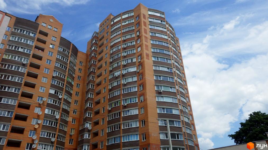 Висотна будівля житлового комплексу з цегляними фасадами на тлі блакитного неба з хмарами. На фасадах видно велику кількість вікон та балконів.