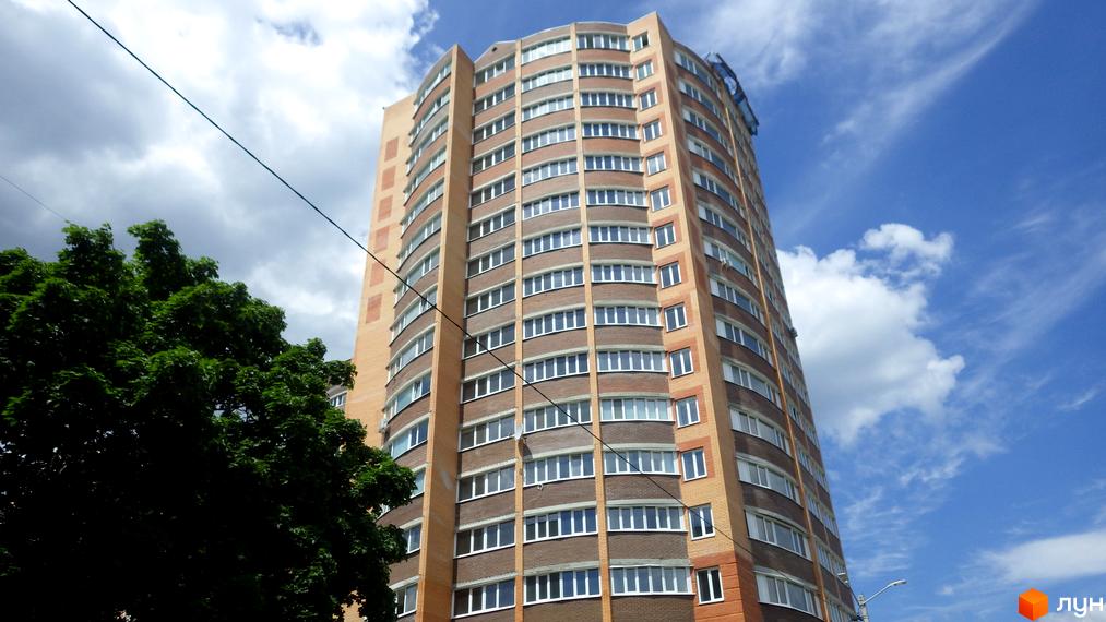 Високий будинок з великою кількістю вікон на тлі блакитного неба з хмарами. Поруч дерева та дроти.