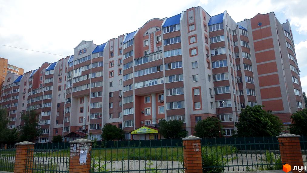 Фасад житлового комплексу на вул. Леваневського, 70, з цегляними та світлими елементами, синім дахом та численними вікнами. На передньому плані зелена огорожа та цегляні стовпчики.