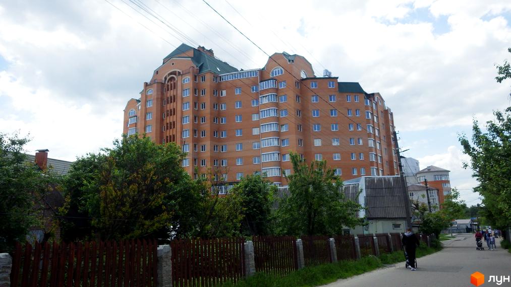 Високий будинок з цегли з великою кількістю вікон. Поруч з будинком ростуть дерева, а також видно дорогу та паркан.