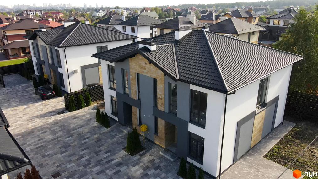 Двоповерховий будинок з темною черепицею та фасадом у світлих тонах. Передня частина будинку оздоблена каменем, перед будинком бруківка та зелені насадження.