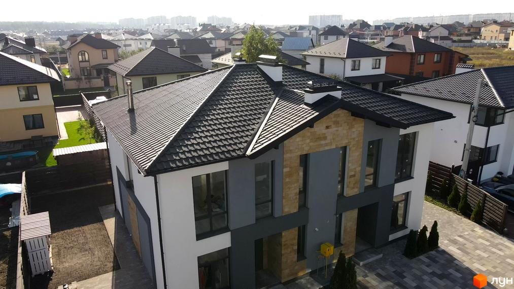 Двоповерховий будинок в Котеджному комплексі Sandstone Comfort з темним дахом та великими вікнами. На території є бруківка та зелені насадження.